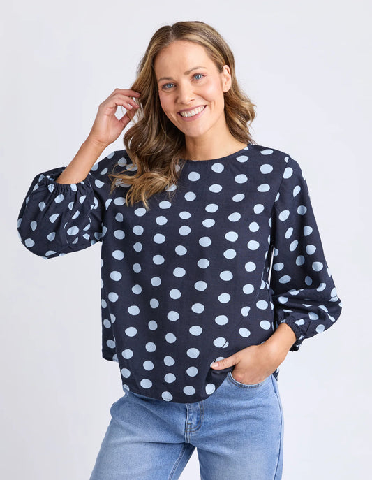 Woman wearing a Elm navy blue blouse with white polka dots and light blue jeans on a plain background