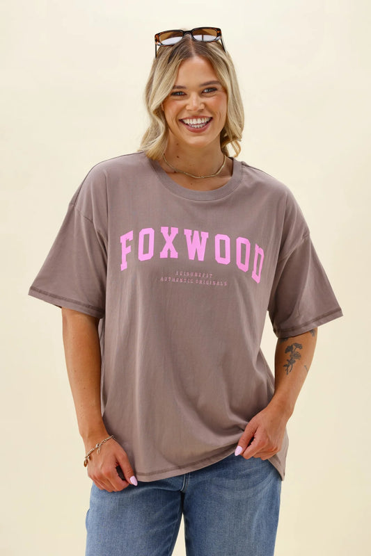 Woman wearing a brown Foxwood interval t-shirt with 'FOXWOOD' printed in pink on a beige background